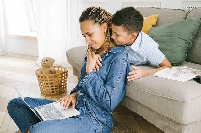 mom with her computer in lap on floor in front of couch with son hugging her